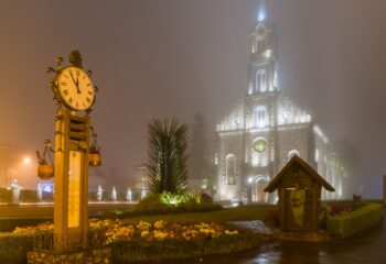 Imagem dos termômetros de Gramado durante a noite, ao fundo, a Igreja Matriz de São Iluminada e desfocada devido a neblina