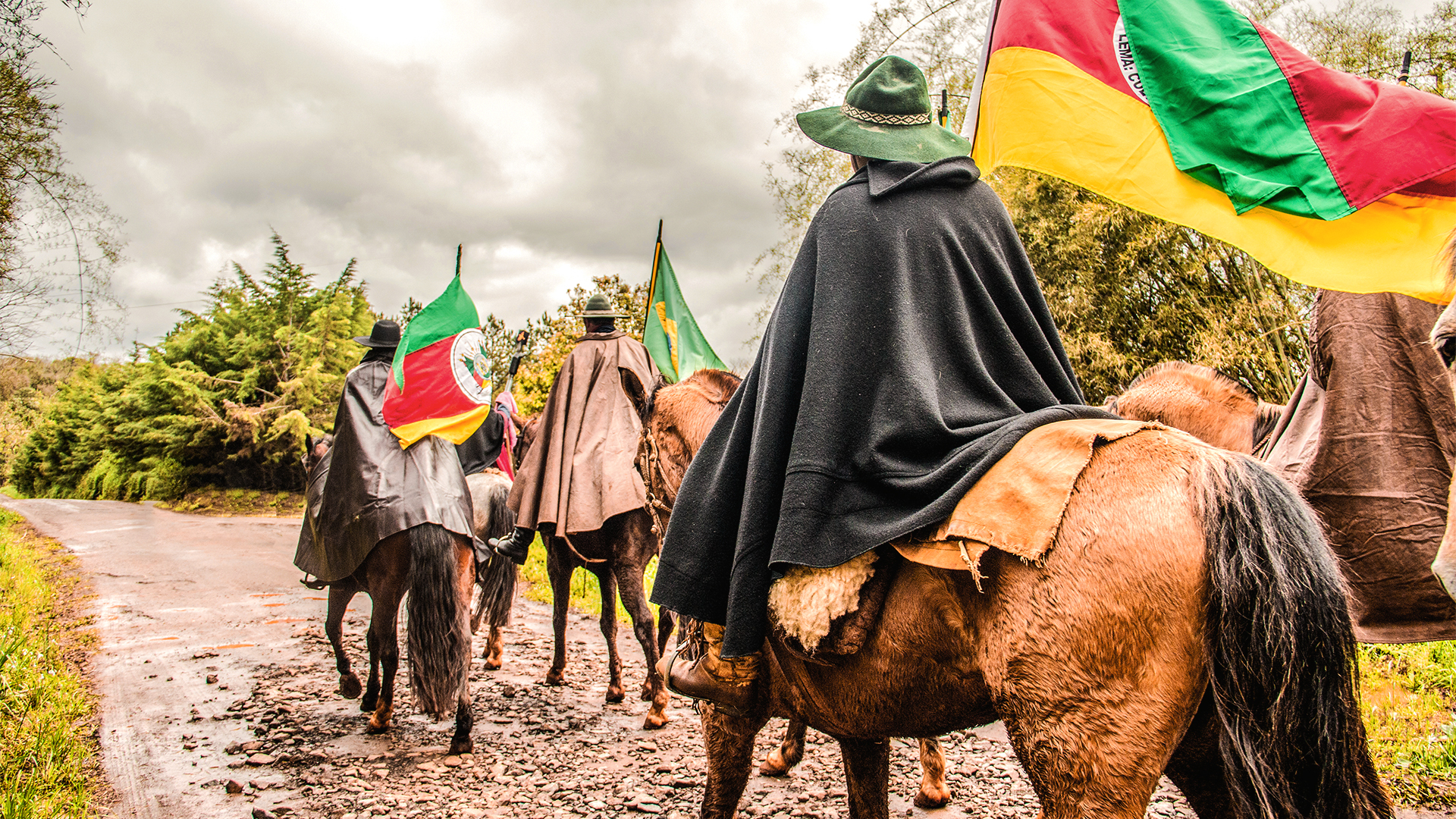Homens trajados com roupas gauchescas montados em cavalos portando bandeiras do Rio Grande do Sul